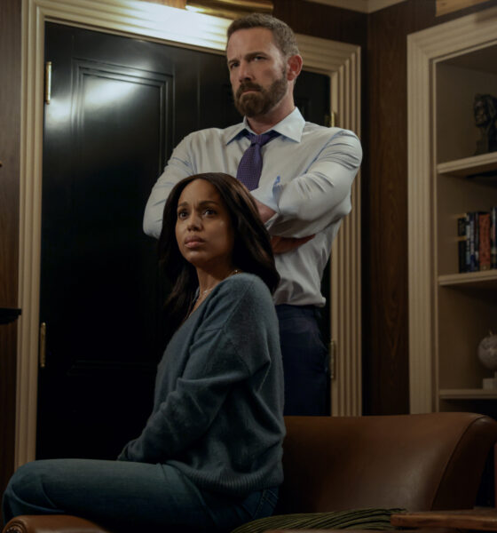 "A woman sits on a leather chair looking concerned, while a man stands behind her with arms crossed in a dimly lit office or library filled with shelves, books, and decorative objects."