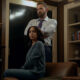 "A woman sits on a leather chair looking concerned, while a man stands behind her with arms crossed in a dimly lit office or library filled with shelves, books, and decorative objects."